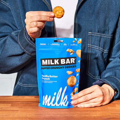 Front view of a bag of vanilla butter crunch crunchies, with a human hand holding up an individual cookie above the bag